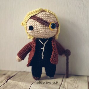 May include: A crocheted doll with blonde hair, wearing a brown jacket, black pants, and an eyepatch. The doll is holding a walking stick. The doll is standing on a wooden surface. The text "@fili.intrecciati" is visible in the bottom right corner of the image.