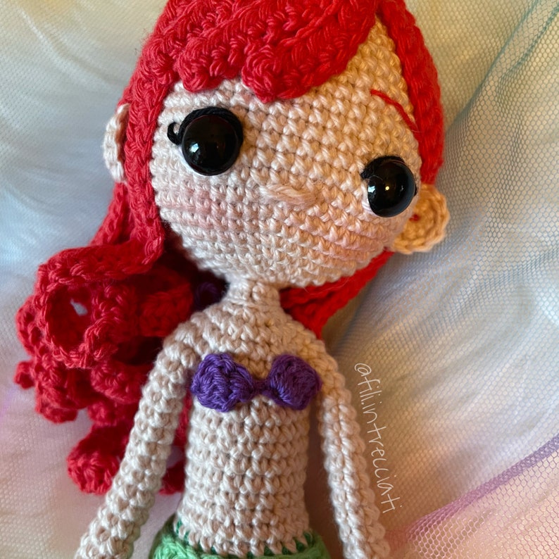 May include: Crocheted Ariel doll with red hair, a purple seashell bra, and big black eyes. The doll is lying on a blue and white tulle fabric.