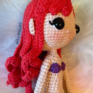 May include: A crocheted doll with red hair, a purple top, and black eyes. The doll is made in a cartoon style and is looking to the right.