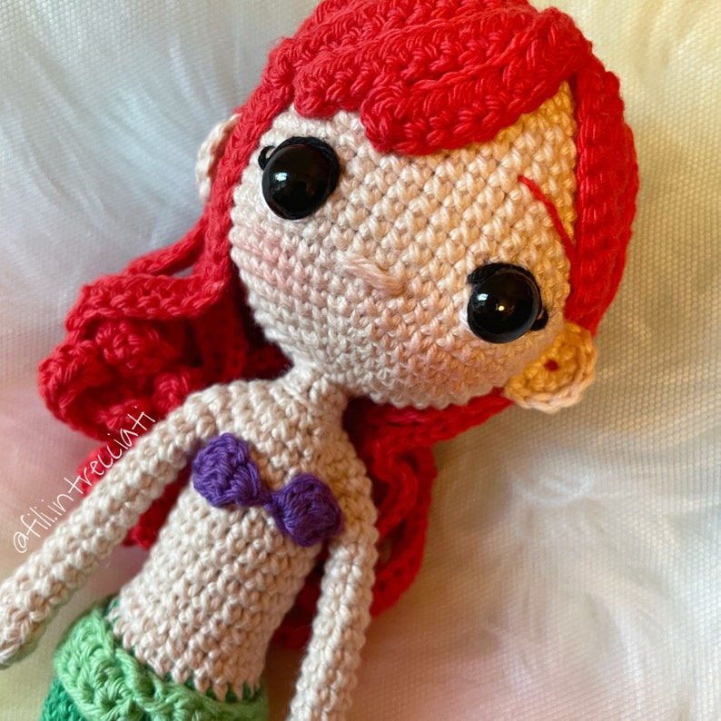 May include: Crocheted amigurumi doll of Ariel, the Little Mermaid, with red hair, a purple seashell bra, and a green mermaid tail. The doll has large black eyes and a sweet expression.