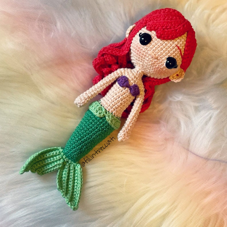 May include: Crocheted mermaid doll with red hair, a purple seashell bra, and a green tail. The doll is lying on a white fluffy surface. The doll's Instagram handle is @filiintrecciati.