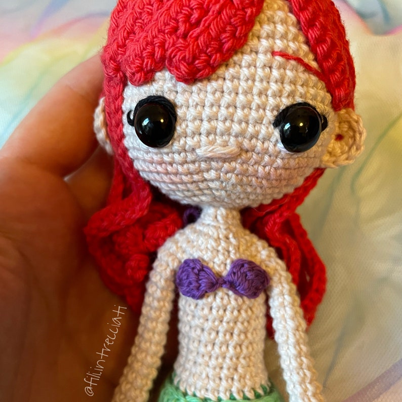 May include: Crocheted doll with red hair, a purple seashell bra, and a green tail. The doll has a white face with black eyes and a pink nose.