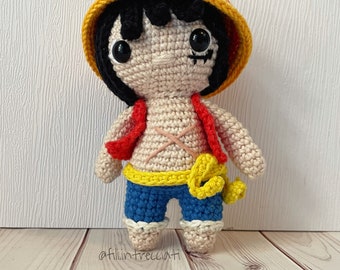 May include: Crocheted toy figure of a character with black hair, wearing a yellow and red hat, a red shirt, a yellow belt, and blue pants. The toy is standing on a wooden surface.