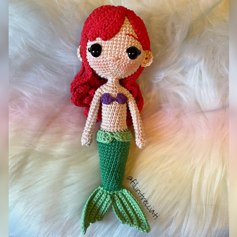 May include: Crocheted mermaid doll with red hair, a purple seashell bra, and a green tail. The doll is sitting on a light green and white fuzzy surface. The text "@filintrecciati" is visible in the bottom right corner of the image.