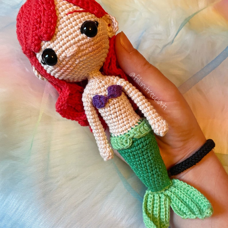 May include: Crocheted mermaid doll with red hair, a purple seashell bra, and a green and white tail. The doll is held in a hand with a black bracelet.
