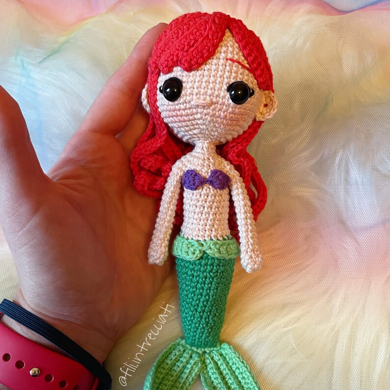 May include: Crocheted mermaid doll with red hair, a purple seashell bra, and a green tail. The doll is holding a seashell in its hand.