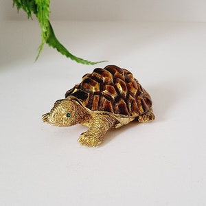 Puede incluir: Una detallada figura de tortuga dorada con caparazón texturizado y ojos verdes. La tortuga está colocada sobre una superficie blanca, con follaje verde en el fondo. La figura es probablemente un objeto decorativo.