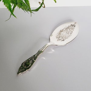 Puede incluir: Un utensilio de servir plateado con un mango adornado y un diseño floral decorativo. El utensilio tiene una cabeza ancha y ovalada. Una planta verde en una maceta con estampado blanco y verde está en el fondo.