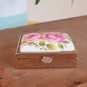 Könnte beinhalten: Kleine, rechteckige silberne Pillendose mit Scharnierdeckel. Der Deckel zeigt ein Blumenmuster mit rosa Rosen und grünen Blättern auf weißem Grund. Die Dose liegt auf einer Holzoberfläche.