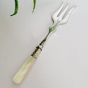 Puede incluir: Un tenedor de servir plateado con mango de nácar. El tenedor tiene tres púas y un diseño decorativo en el mango. El mango es de color blanco cremoso. El tenedor se utiliza probablemente para servir aperitivos o postres.