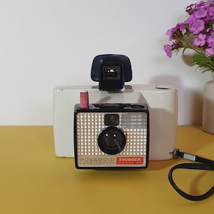 May include: A vintage Polaroid Swinger Land Camera Model 30 with a black viewfinder and a red button. The camera has a silver and black face with the words "POLAROID SWINGER LAND CAMERA" printed on it. A black wrist strap is attached. A small vase of flowers is in the background.