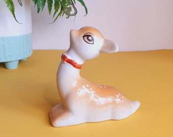 May include: A ceramic deer figurine, white with tan spots, and a red collar. The deer is in a seated pose. A potted fern is in the background. The figurine is likely a decorative item.