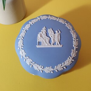 Könnte beinhalten: Hellblaue Keramik-Schmuckdose mit weißem Reliefdesign von Figuren und einem Lorbeerkranz-Rand. Die Dose steht auf einer gelben Oberfläche, im Hintergrund eine kleine weiße Vase mit rosa und weißen Blumen.