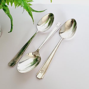 Puede incluir: Tres cucharas plateadas con superficies reflectantes están dispuestas sobre una superficie blanca. Las cucharas tienen mangos alargados y cuencos redondeados. Una cuchara tiene un diseño floral en el mango, mientras que otra tiene un pequeño emblema. Una planta verde en una maceta estampada blanca y gris está en el fondo.