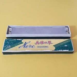 May include: A vintage harmonica in its original box. The harmonica is silver and rests in a white-lined box. The box is green and yellow with the word "Hero" and Chinese characters printed on it. The harmonica is a musical instrument.