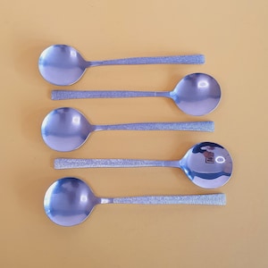 May include: Five stainless steel spoons with round bowls and textured handles are arranged on a light orange surface. The spoons are reflective, and the handles have a linear pattern.