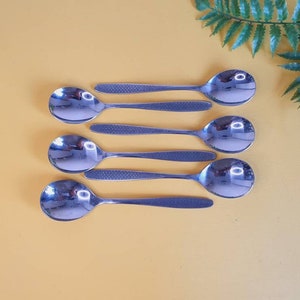 May include: Six stainless steel spoons on a yellow surface. The spoons have a reflective, polished finish with textured handles. A fern leaf is in the background. These spoons are designed for serving. The spoons are made of stainless steel.