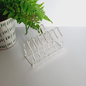 Könnte beinhalten: Ein silberfarbener Metall-Toastständer mit geometrischem Design. Der Ständer hat sechs Schlitze und einen Griff. Im Hintergrund befindet sich ein Topf mit Farn.