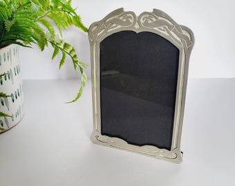 May include: A silver-coloured picture frame with an ornate, Art Nouveau-inspired design. The frame has a black interior and is standing upright. A green plant in a white and green patterned pot is in the background.