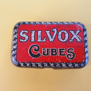 Puede incluir: Lata rectangular vintage con fondo rojo y texto "SILVOX CUBES" en blanco y negro. La lata tiene un aspecto desgastado, con un borde a cuadros blanco y negro.