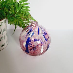 Puede incluir: Un jarrón de vidrio redondo con un diseño moteado en tonos rosa, azul y blanco. El jarrón tiene un cuello estrecho y probablemente se utiliza con fines decorativos. El diseño del jarrón recuerda a una pintura de acuarela.