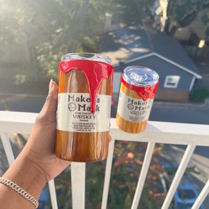 May include: Two stainless steel tumblers with a wood grain design and red dripping paint. The tumblers are labeled "Maker's Mark Whiskey" and feature the brand's logo.