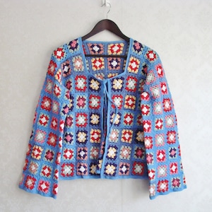 Puede incluir: Un cárdigan de crochet azul con un frente de corbata y patrones cuadrados coloridos. El cárdigan tiene mangas largas y un ajuste holgado.