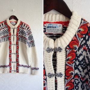 Puede incluir: Un cárdigan blanco con un diseño estampado en rojo, azul y blanco. El cárdigan tiene botones de metal plateado y una etiqueta que dice "Devold, Made in Norway, 100% Pure New Wool".