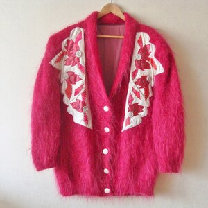 May include: A fuzzy pink cardigan with white and red floral embroidery on the front. The cardigan has a V-neck and button closure.