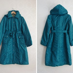 Può includere: Un cappotto trapuntato blu turchese con cappuccio e cintura. Il cappotto ha una chiusura con bottoni e due tasche.