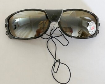 May include: Black sunglasses with a mirrored lens and a black strap. The sunglasses have a black leather strap that goes over the top of the bridge of the nose. The strap has a small metal buckle. The sunglasses have a small round sticker on the right lens that says "All Weather Lens, 100% UV Protection".