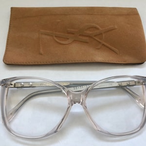 Puede incluir: Gafas de vista con montura de plástico transparente, puente y patillas de metal plateado. Las patillas están etiquetadas con "Yves Saint Laurent" y "58-16-140". Las gafas vienen con una funda de ante marrón.