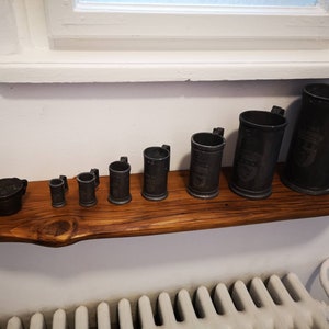 May include: A set of eight pewter mugs of varying sizes, arranged in order of size on a wooden shelf. The mugs are all dark grey in colour and have a slightly aged appearance.