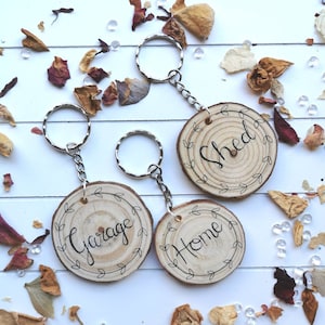 May include: Four wooden keychains with silver key rings. Each keychain is a circular wood slice with a black script word: "Garage", "Home", and "Shed". The words are surrounded by a decorative leaf design. The background has dried flowers and clear beads.