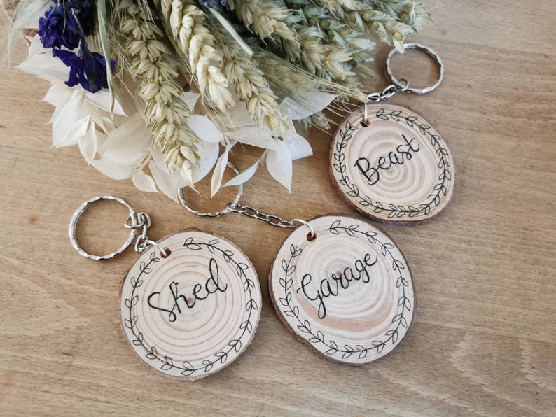 May include: Wooden keychains with silver key rings. Each keychain is a light brown, oval-shaped wood slice with a black handwritten word: "Beast", "Shed", and "Garage". Each keychain has a black leaf border.