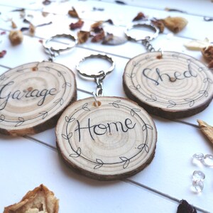 May include: Three wooden keychains with silver rings. Each keychain is a circular wood slice with the words "Garage", "Home", and "Shed" in black script. The words are surrounded by a leaf design. The background has scattered dried flowers.