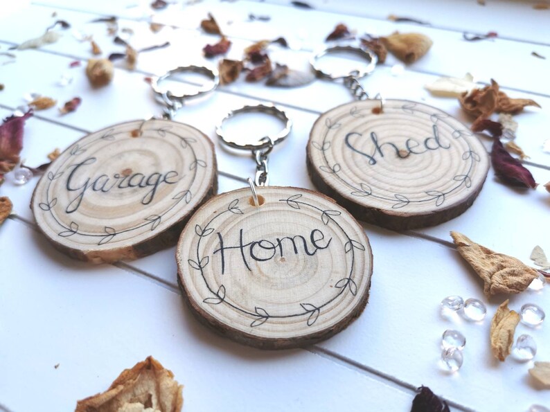 May include: Three wooden keychains with silver rings. Each keychain is a circular wood slice with the words "Garage", "Home", and "Shed" in black script. The words are surrounded by a leaf design. The background has scattered dried flowers.