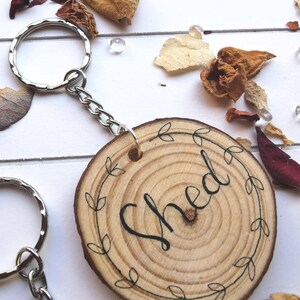 May include: Wooden keychain with the word "Shed" in black script, surrounded by a leaf design. The keychain is attached to a silver metal ring and chain. The wood slice has visible growth rings.