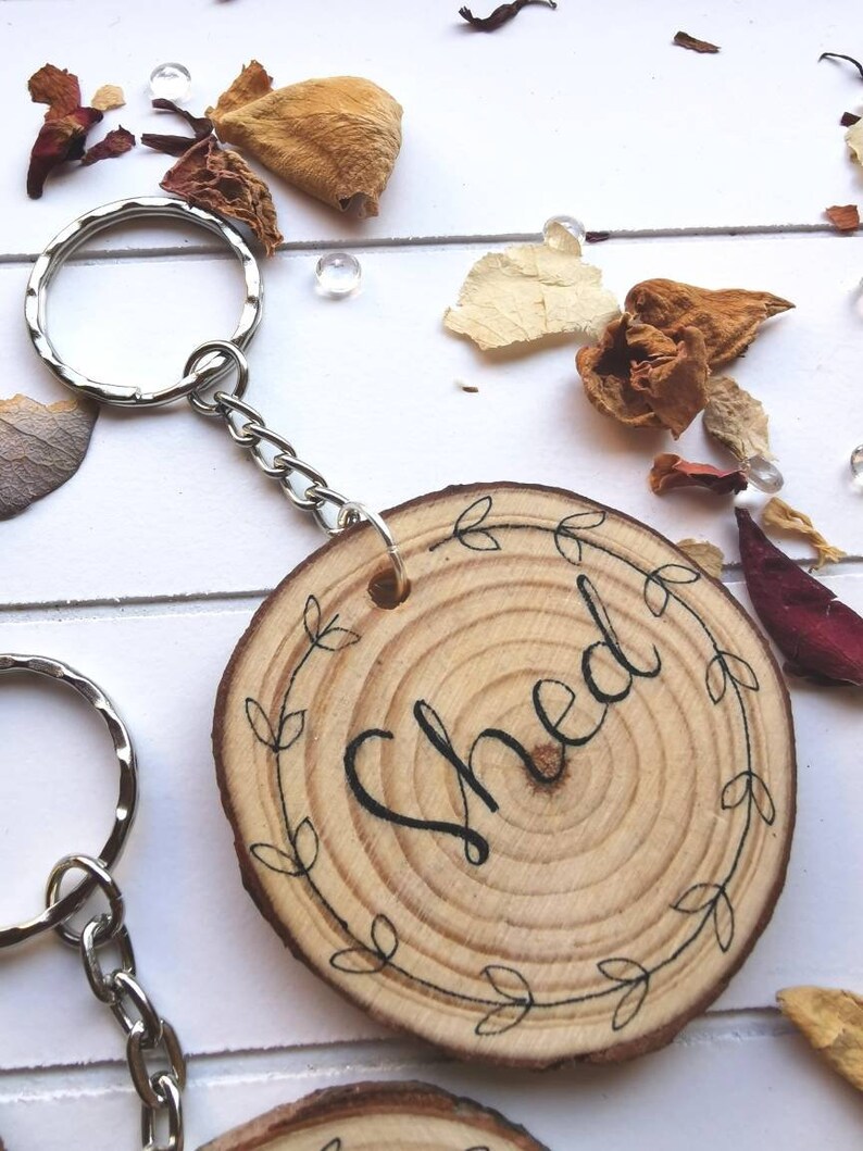 May include: Wooden keychain with the word "Shed" in black script, surrounded by a leaf design. The keychain is attached to a silver metal ring and chain. The wood slice has visible growth rings.