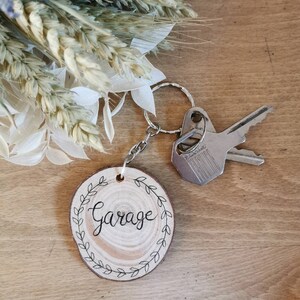 May include: A wooden garage keychain with a silver chain and keyring. The round wooden tag has the word "Garage" in black script, surrounded by a leaf design. Two keys are attached to the keyring. The background includes dried flowers.