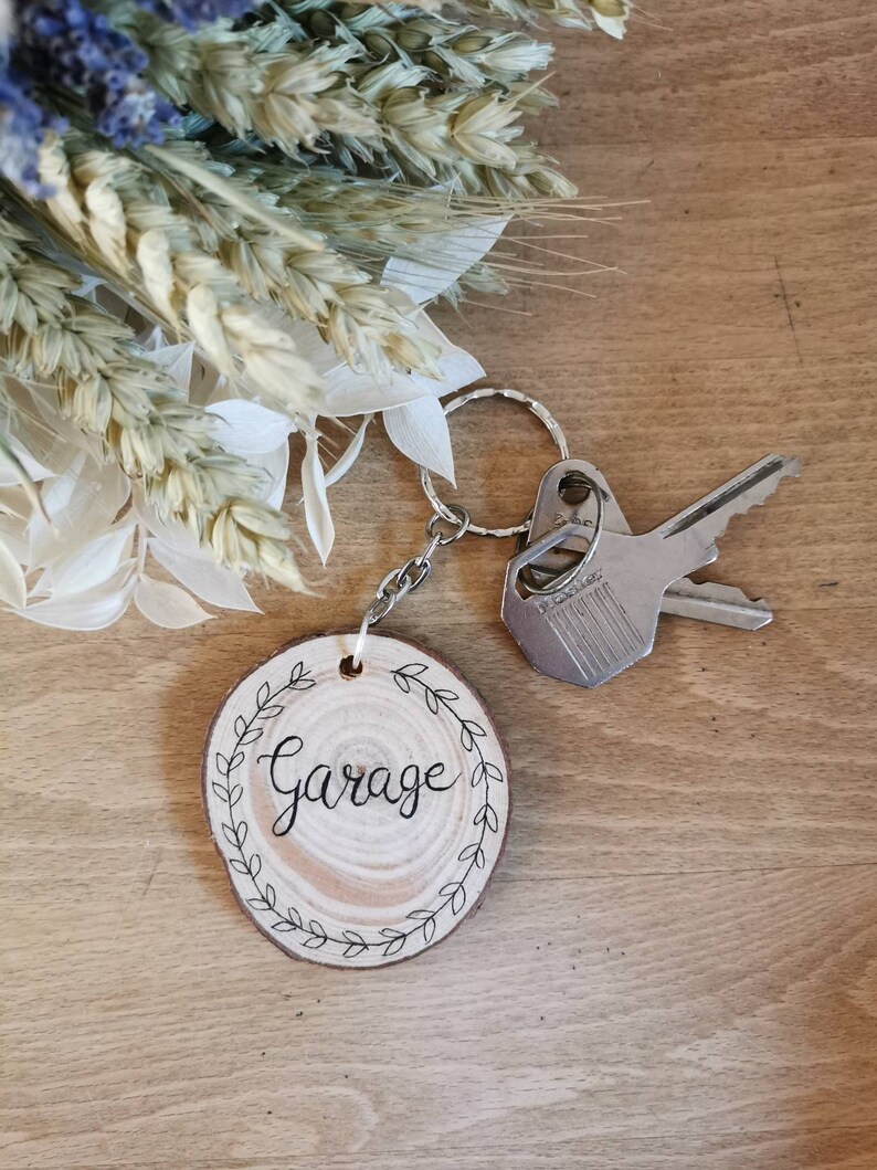 May include: A wooden garage keychain with a silver chain and keyring. The round wooden tag has the word "Garage" in black script, surrounded by a leaf design. Two keys are attached to the keyring. The background includes dried flowers.
