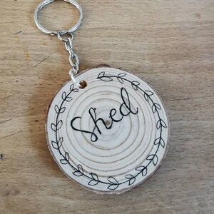 May include: A wooden keychain with the word "Shed" in black script. The round wooden piece is decorated with a black leaf wreath. The keychain has a silver metal ring and chain.