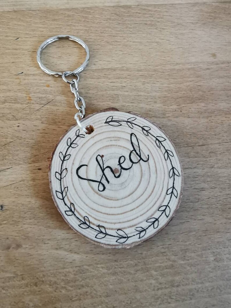 May include: A wooden keychain with the word "Shed" in black script. The round wooden piece is decorated with a black leaf wreath. The keychain has a silver metal ring and chain.
