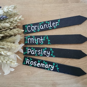 May include: Four black wooden garden markers with white lettering and green vine accents. The markers read "Coriander", "Mint", "Parsley", and "Rosemary". A bouquet of dried flowers is in the background.