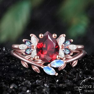 May include: A rose gold ring featuring a central pear-cut deep red garnet stone, flanked by marquise-cut white and iridescent blue stones arranged in a leaf-like pattern. The band is shaped like a vine with small leaves. Text reads LOVE HSW JEWELRY ART.
