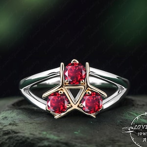 Könnte beinhalten: Ein silberner Ring mit drei leuchtend roten Edelsteinen, die in einem goldenen Dreieck gefasst sind. Der Ring hat ein geteiltes Schaftdesign und ist vor einem dunkelgrünen Hintergrund platziert. Der Ring ist ein Schmuckstück.
