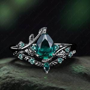 Könnte beinhalten: Silbernes Ring-Set mit einem birnenförmigen, smaragdgrünen Edelstein als Mittelpunkt. Der Ring ist mit kleineren, runden, teal-farbenen Edelsteinen und filigranen, blattartigen Details verziert. Das Band hat ein aufwendiges, von der Natur inspiriertes Design.