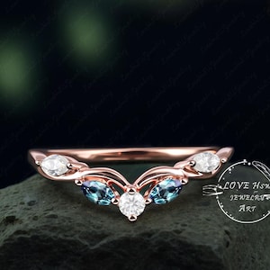 May include: A rose gold wedding band with a delicate, curved design. The band features three blue gemstones and a larger, round, white gemstone in the center. The band is set on a dark, textured surface. "LOVE HOW JEWELRY ART" is written in white text on a black background.
