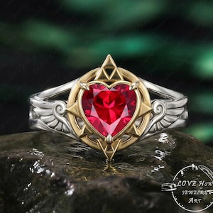 Könnte beinhalten: Ein silberner Ring mit einem goldfarbenen herzförmigen Edelstein. Der Edelstein hat eine leuchtend rote Farbe und ist in einen goldenen Rahmen mit Kompassdesign gefasst. Der Ring hat flügelartige Details an den Seiten. Der Ring befindet sich auf einer dunklen, strukturierten Oberfläche.