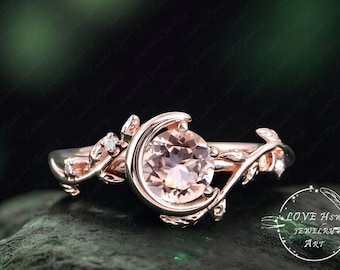 May include: Rose gold ring featuring a round, peach-colored gemstone set within a crescent moon design. The ring is adorned with delicate leaf and vine details, and a small diamond accent. The ring is set against a dark green background.